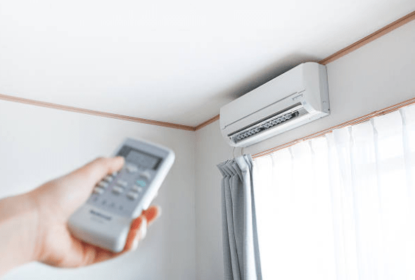 A person holds a remote control pointed toward a wall-mounted split system air conditioner unit in a room.