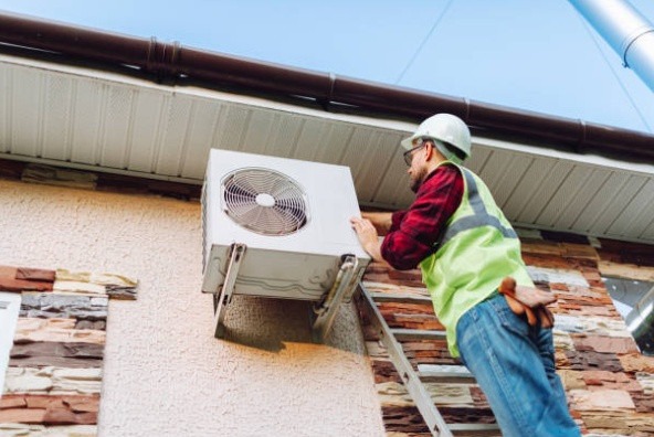 A technician wearing a hard hat and safety vest stands on a ladder to install an outdoor air conditioning condenser unit on the side of a building.