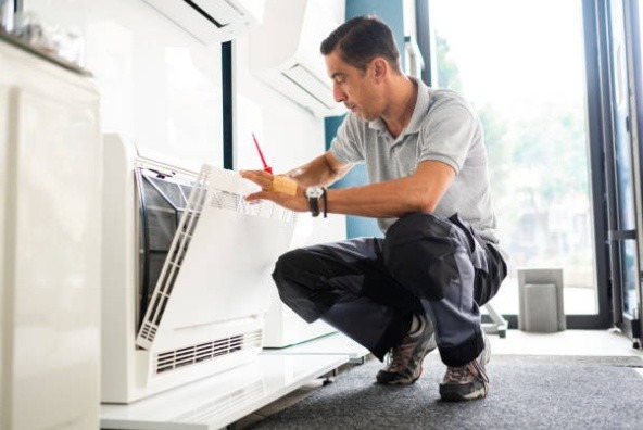 A technician kneels on the floor while using a tool to open and inspect the front panel of an indoor air conditioning unit.