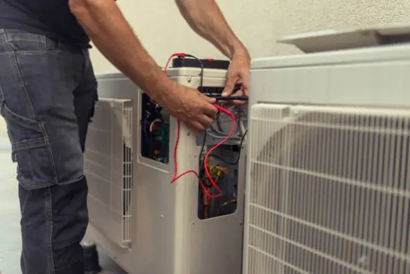 A service technician uses a multimeter with red and black leads to test the electrical components inside an outdoor air conditioning unit.