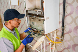 Technician in a hi-vis vest and safety glasses inspecting the internal wiring and components of an open wall-mounted boiler