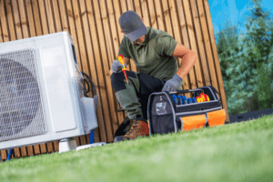 Technician in a cap kneeling beside an outdoor air conditioning unit, selecting a screwdriver from an open toolkit on the grass