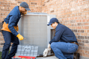 Two HVAC technicians working together on an outdoor condenser unit against a brick wall, one holding pliers and the other opening a toolbox