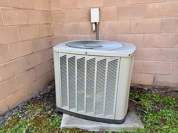 Older central air conditioner condenser on a concrete pad beside a tan cinder block wall