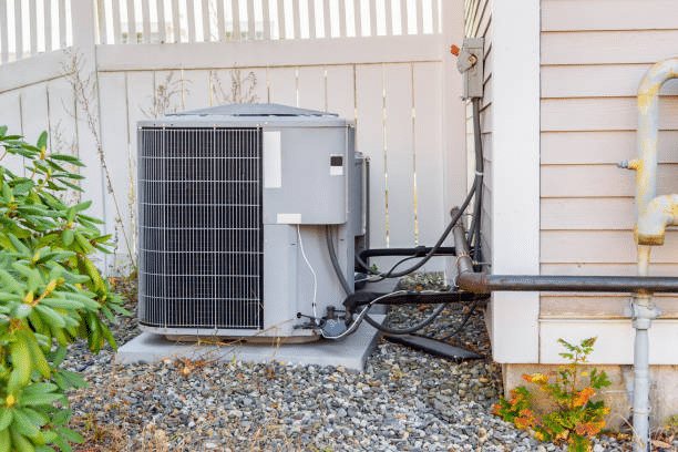 Outdoor HVAC condenser unit on a pad in a gravel bed with refrigerant lines and pipes along the house siding