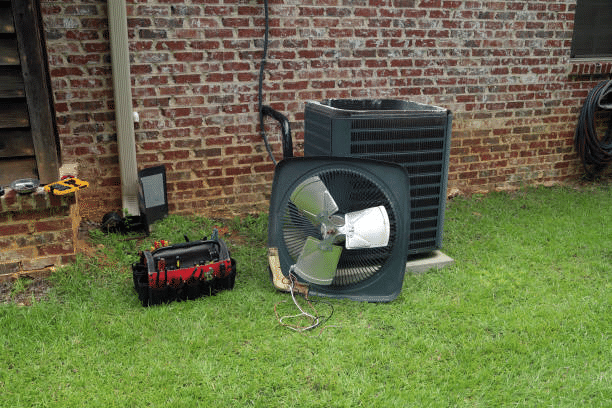 AC condenser unit with its fan removed and a toolbag on the lawn during repair beside a brick wall