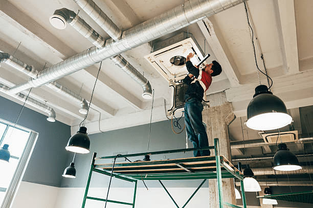 Technician on a scaffold installing a ceiling cassette air conditioner beside exposed ductwork and pendant lights