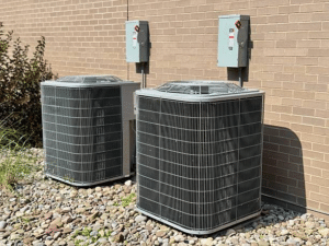 Two outdoor AC condenser units on river rock beside a tan brick wall with electrical disconnect boxes
