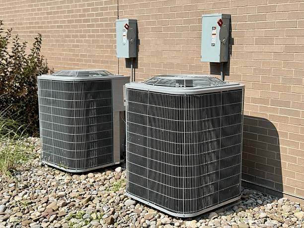 Two outdoor AC condenser units on river rock beside a tan brick wall with electrical disconnect boxes